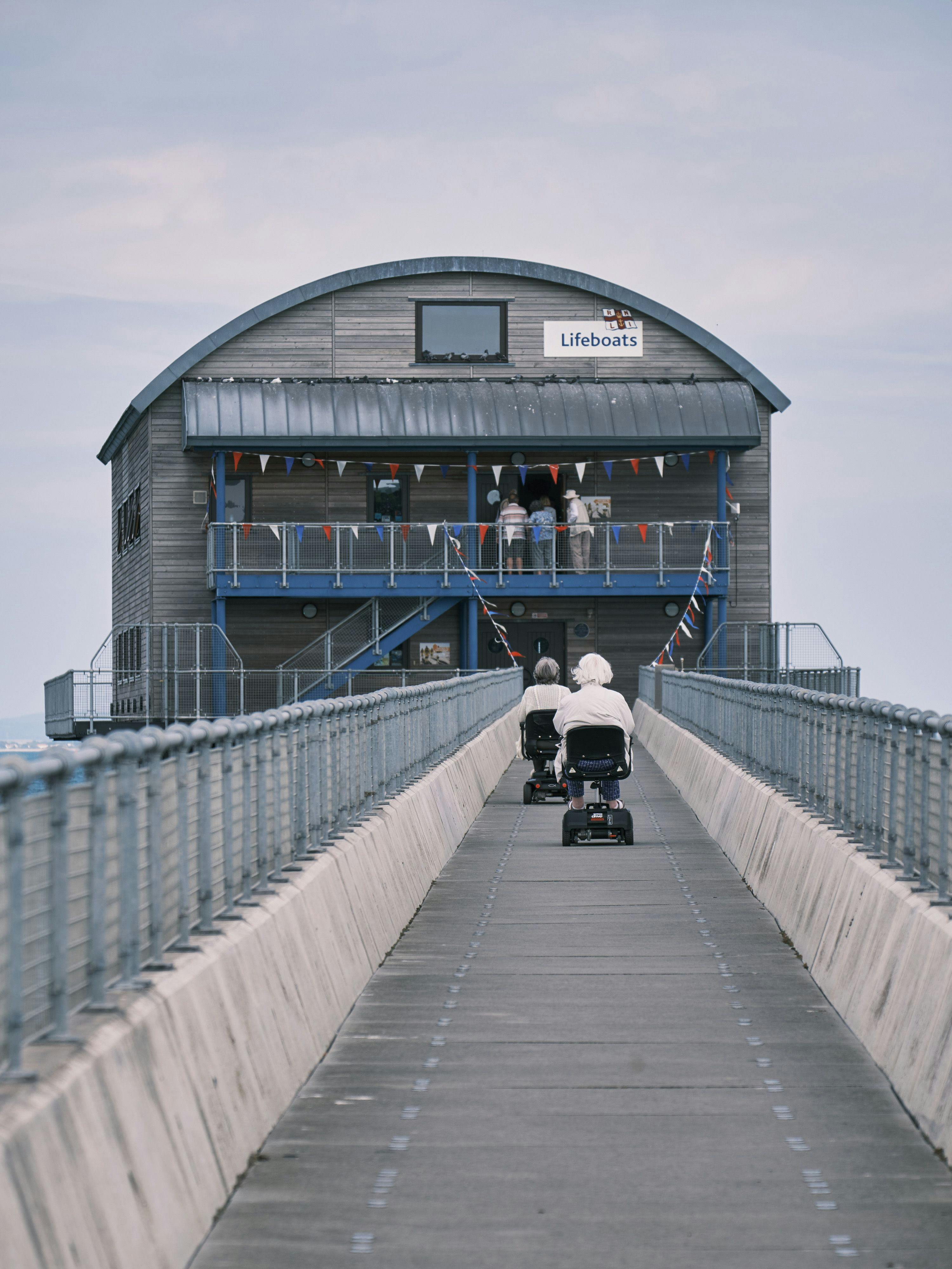 Two mobility scooters drive up a pier towards a lifeboat station