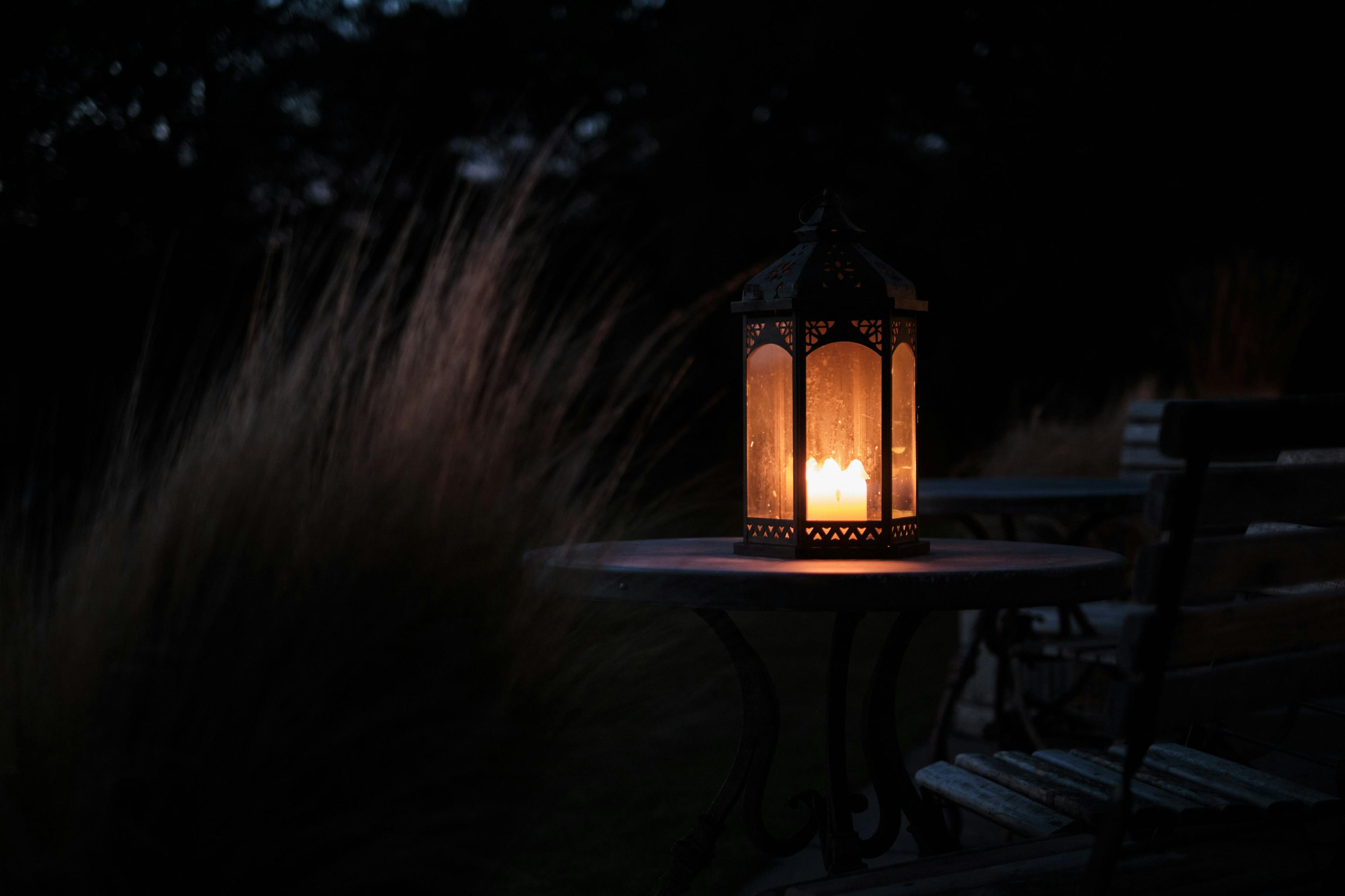A candle lit lantern on a table barely illuminates some long grass. 