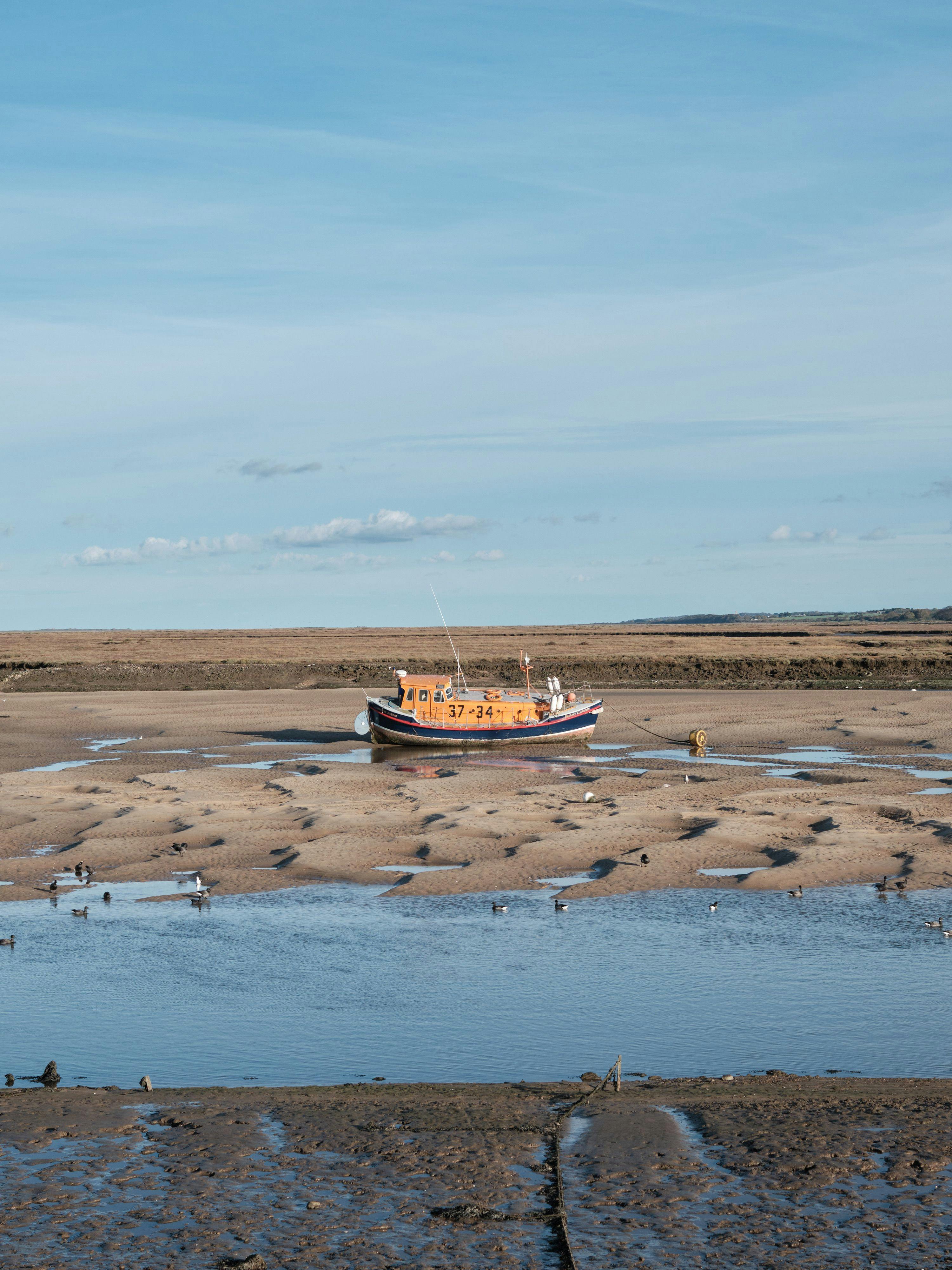 A lifeboat resting on the sand as the tide has gone out. The orange of the boat contrasts with the blue of the sky and yellowy sand